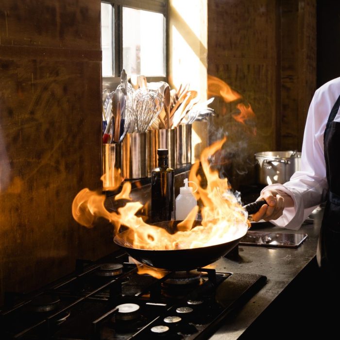 Side view of a Caucasian woman and an African man at a cookery class, frying vegetables on a frying pan, flaming